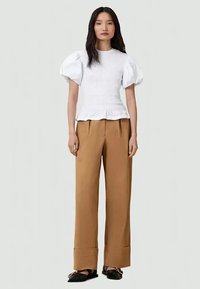 Top bianco arricciato con maniche a sbuffo, pantaloni larghi marroni e scarpe slip-on nere. L'outfit presenta una finitura testurizzata e una vestibilità sartoriale.