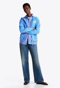 Sudadera azul clara con bolsillos frontales, llevada sobre una camiseta a rayas azul y blanca, combinada con jeans de pierna ancha y zapatos marrones.