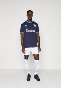 Maillot de football de la marine avec des accents turquoise, comportant un logo et un texte de sponsor. Porté avec un short blanc et des chaussettes blanches arrivant aux genoux, des crampons noirs.