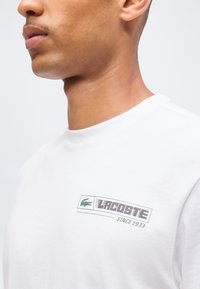 Άνδρας που φοράει λευκό πουκάμισο Lacoste με πράσινο λογότυπο κροκοδείλου και κείμενο "Lacoste Since 1933" στο στήθος.