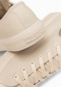 Baskets en toile beige avec lacets et semelle texturée portant le nom de la marque embossé "OMERE" sur le talon.