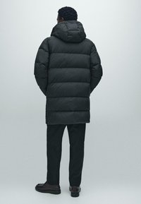 Uomo in piedi di spalle, con indosso un lungo giubbotto in stile piumino nero con cappuccio, pantaloni neri e scarpe scure su uno sfondo grigio.