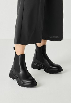Person wearing black leather Chelsea boots with thick soles and black wide-leg pants standing on a light floor.
