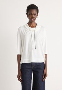 Witte lange mouw top met capuchon en een gestructureerde afwerking. Voorzien van een trekkoord met zilveren uiteinden. Gekleed met donkerblauwe wijde jeans.