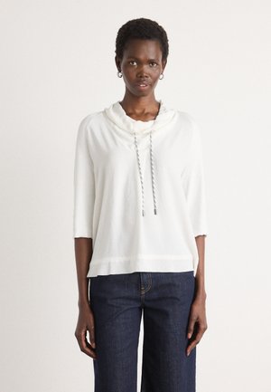 Witte lange mouw top met capuchon en een gestructureerde afwerking. Voorzien van een trekkoord met zilveren uiteinden. Gekleed met donkerblauwe wijde jeans.