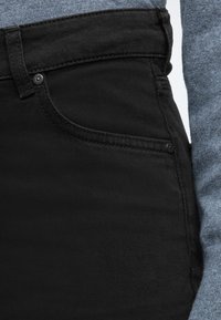 Čierne rifľové džínsy s hladkou textúrou, úzkym strihom a jemným prešívaním. Majú predné vrecko a zapínanie na gombík v páse.