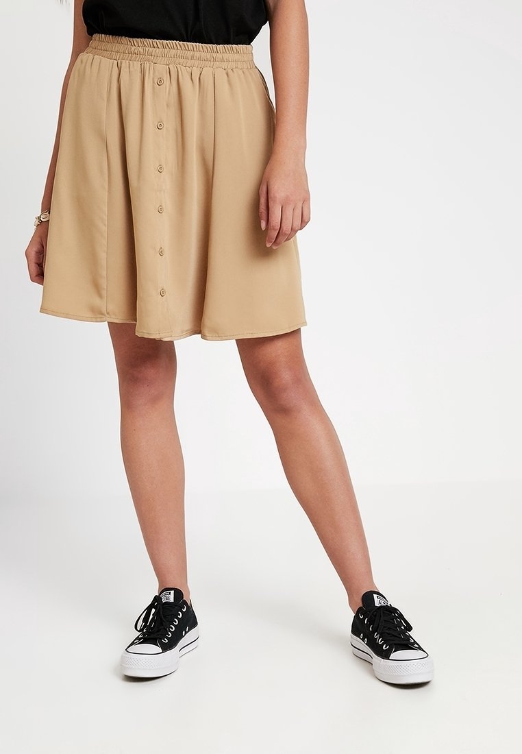 Femme portant une jupe beige boutonnée sur le devant avec une taille élastique et des baskets noires, debout sur un fond blanc.