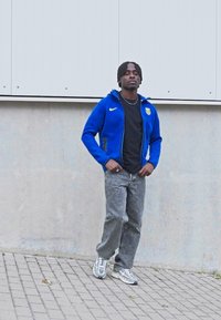 Μπλε zip-up hoodie της Nike με λογότυπο, μαύρη μπλούζα, γκρι τζιν και σε sneakers με σχέδια. Στέκεται σε μια τοιχοποιία από τούβλα μπροστά σε ένα σκυρόδεμα τοίχο.