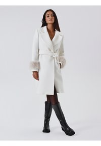 Weißer Trenchcoat mit tailliertem Design, einem Gürtel und plüschigen Fellmanschetten, aus glattem, strukturiertem Stoff. Schwarze knielange Stiefel.