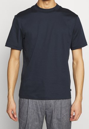 Homme portant une chemise ajustée à manches courtes bleu marine foncé et un pantalon gris à carreaux, debout les bras détendus le long du corps.