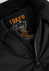 Czarna kurtka z puszystym kołnierzem, z naszywką logo pantery z napisem "TOKYO" i "Japonia" w kolorze pomarańczowym. Zamek błyskawiczny z przodu.