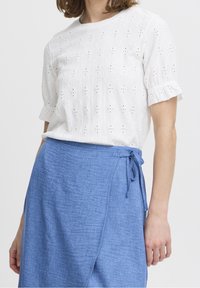 Blusa bianca con occhielli e maniche corte, caratterizzata da intricati motivi a taglio. Abbinata a una gonna avvolgente leggera di colore blu chiaro, annodata in vita.