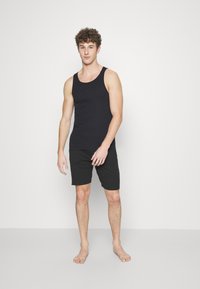 Ein Mann steht und trägt ein schwarzes Tanktop und dunkle Shorts. Das Tanktop ist tailliert und hat breite Träger, und der Stoff wirkt glatt.