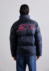 Doudoune bleue marine avec une texture volumineuse. Présente un graphique rose à l'arrière d'un chat en train de sauter. Assortie à un jean en denim bleu.