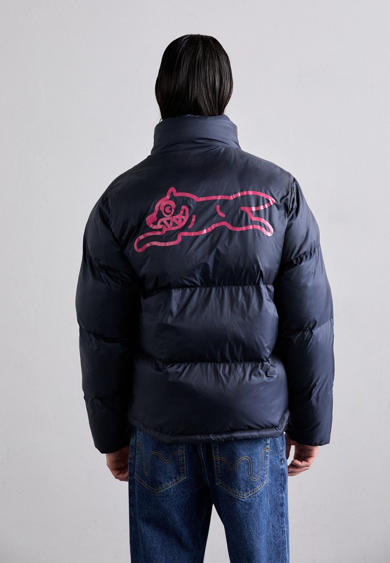 Doudoune bleue marine avec une texture volumineuse. Présente un graphique rose à l'arrière d'un chat en train de sauter. Assortie à un jean en denim bleu.
