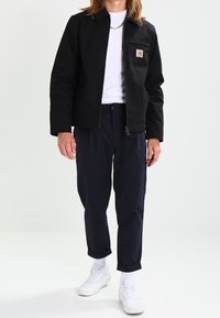 Černá plátěná bunda se zipem a náprsní kapsou, doplněná tmavými kalhotami a bílými teniskami. Pod tím bílý tričko.