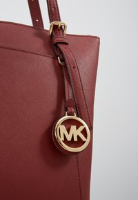 Gros plan sur la lanière rouge en cuir d'un sac à main avec une boucle réglable et un portant un pendentif rond doré avec le logo MK.