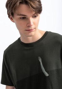 Donkergroen katoenen t-shirt met een ronde halslijn, voorzien van een afbeelding van een opgestoken hand aan de voorkant, gedeeltelijk verborgen door een schaduw.