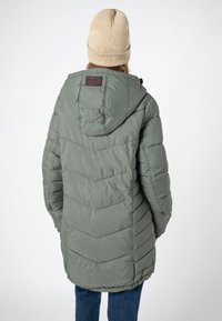 Persoon in een groene gewatteerde jas met capuchon en een beige muts, gezien van achteren, staand tegen een effen lichte achtergrond.