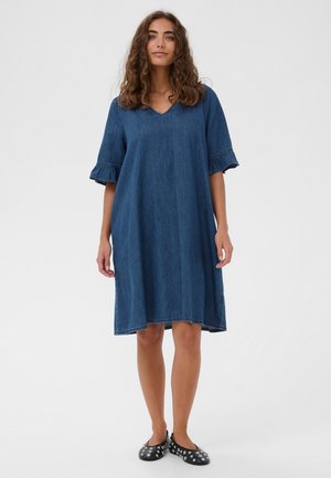 Göndör hajú nő, laza kék farmerruhában, fodros rövid ujjakkal és fekete szegecsekkel díszített lapos cipőben, fehér háttér előtt áll.