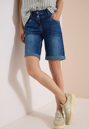 Personne portant un short en jean bleu retroussé, une chemise rayée et des baskets beiges, debout avec une main dans la poche, face à un mur clair.