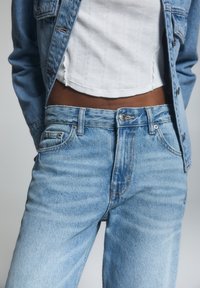 Persona vestida con jeans de mezclilla azul claro y chaqueta, con una blusa corta blanca, de pie contra un fondo gris neutro.