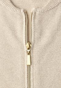 Primo piano di un capo a maglia beige con una cerniera dorata parzialmente aperta al centro.