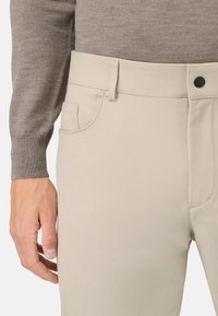 Lys beige bukser med en glat tekstur, detaljer ved forlommerne og en knaplukning i taljen, båret med en grå sweater.