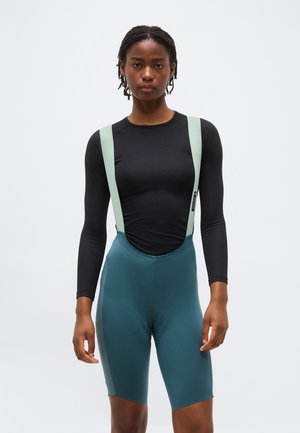 Mujer con camiseta negra de manga larga y pantalones cortos de ciclismo tipo peto en verde azulado con tirantes verde claro, de pie frente a un fondo blanco liso.