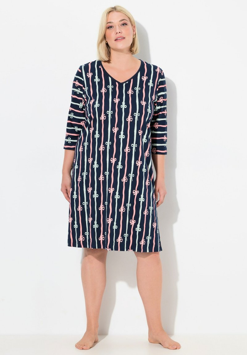 Donna in piedi a piedi nudi che indossa un abito navy lungo fino al ginocchio con strisce verticali rosa e bianche e motivi a fiocco, con maniche a tre quarti.
