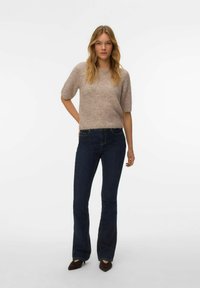 Femme aux longs cheveux blonds portant un pull en maille beige à manches courtes, un jean bootcut bleu foncé et des chaussures pointues marron foncé sur fond blanc.