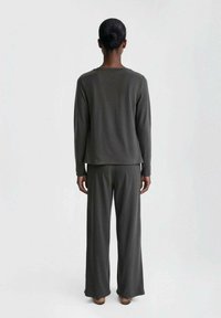 Person with dark hair in a bun wearing a dark gray long-sleeve top and wide-leg pants, standing facing a plain white background.
