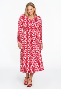 Rotes und weißes langärmliges Kleid mit Wickeldesign, das ein Blattmuster aufweist, einer gebundenen Taille und einem fließenden Rock, kombiniert mit roten Riemchensandalen.