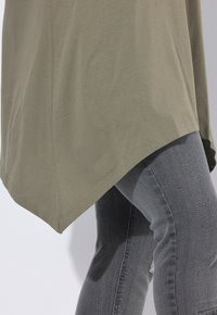 Tunika de manga larga verde oliva con dobladillo curvado, combinada con jeans oscuros de corte slim con un ligero desgarro en la rodilla.