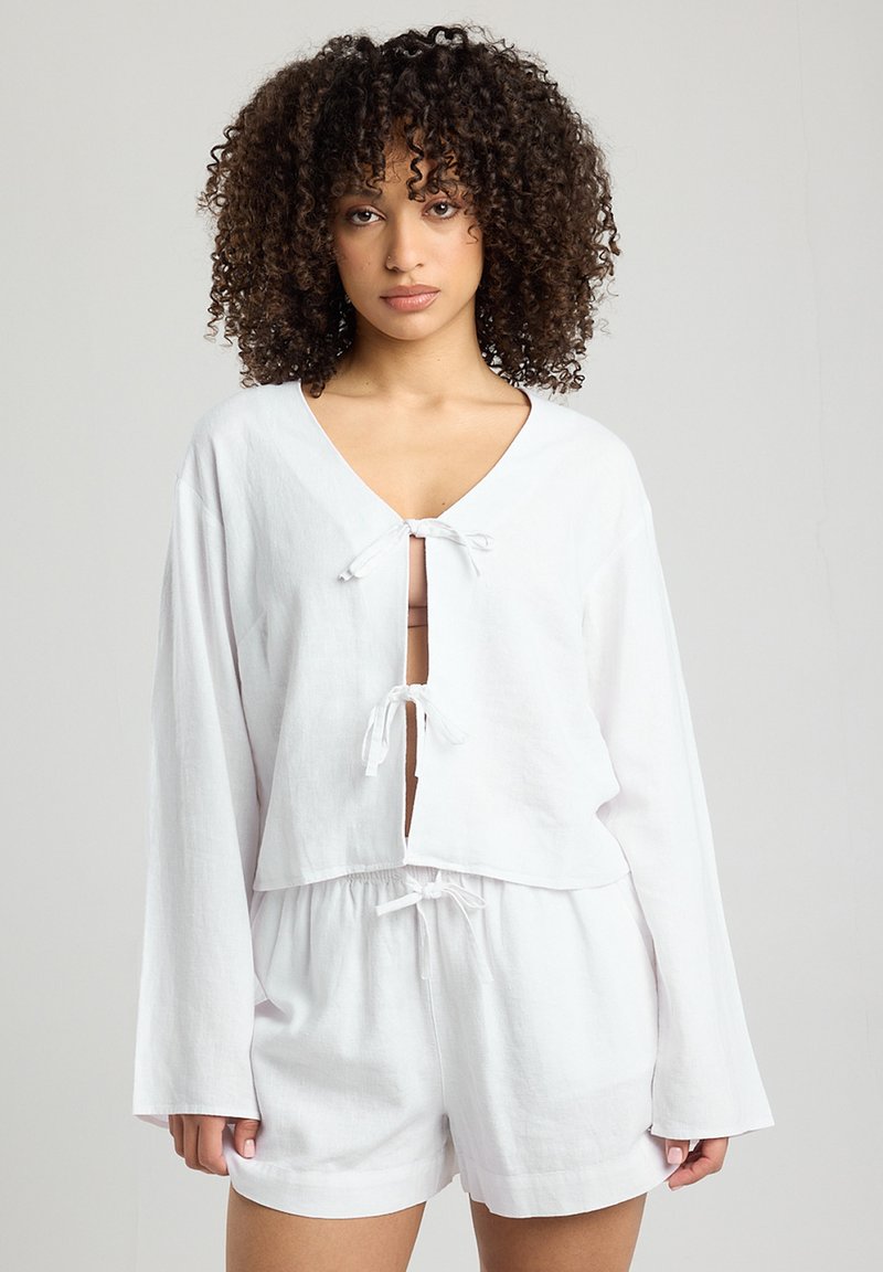 Top corto in lino bianco con maniche lunghe, scollo a V e chiusura con fiocco frontale. Abbinato a pantaloni corti bianchi coordinati con vita elastica.