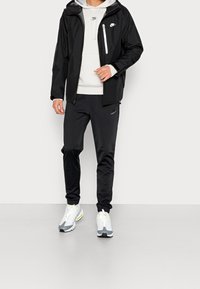 Svart Nike träningsjacka med huva, vit ficka med dragkedja, tillsammans med svarta byxor med logotyp. Grå och vita sneakers med gula detaljer.