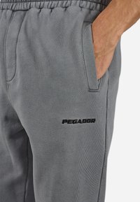 Pantaloni de trening gri, fabricați dintr-un material moale, cu un brâu elastic, buzunare laterale și un logo negru "PEGADOR" brodat pe coapsa stângă.