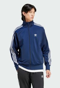 Veste bleu marine avec un col montant, design à trois rayures blanches sur les manches, fermeture éclair à l'avant et petit logo Trefoil sur la poitrine.