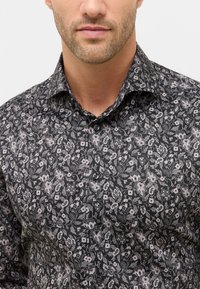 Man in een zwart knoopshirt met een grijs bloemen- en paisleypatroon, met zijn onderste gezicht en hals zichtbaar tegen een effen achtergrond.