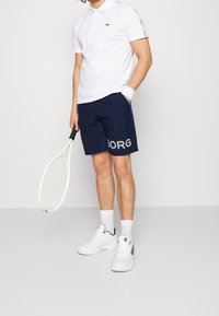 Vit pikétröja, marinblå shorts med "BORG" tryckt i grått, vita atletiska skor, håller en tennisracket, handledsband på vänster handled.