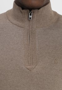 Beige Halbzip-Pullover mit geripptem Kragen, strukturierter Strickmuster und kleinem Logo unten rechts.