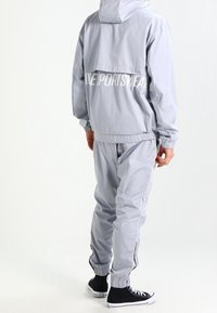 Lichtgrijze trainingsoutfit van glad materiaal met een capuchonjack, elastische manchetten en de tekst "LIVE SPORTSWEAR" op de achterkant.