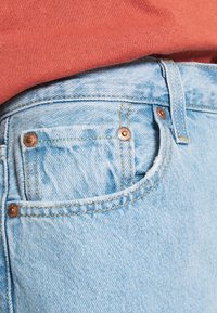 Jasnoniebieskie dżinsy z widocznym przeszyciem, miedzianymi nitami i małą przednią kieszonką, zestawione z czerwono-pomarańczową bluzką materiałową.