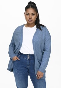 Világoskék kardigán puha kötött anyagból, laza szabással, nyitott elülső résszel és bordázott mandzsettákkal. Fehér inggel és farmernadrággal viselve.