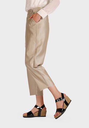 Sandali wedge in vernice nera con suole simili al sughero e fibbie regolabili, abbinati a pantaloni beige chiaro a gamba larga.