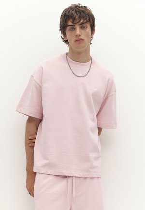 Jeune homme portant un t-shirt oversize rose clair et un short assorti, avec un collier en chaîne argentée, sur un fond blanc uni.