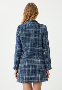 Kobieta z długimi blond włosami, ubrana w niebieską i białą w kratkę sukienkę z długim rękawem, stojąca tyłem na tle gładkiego białego tła.