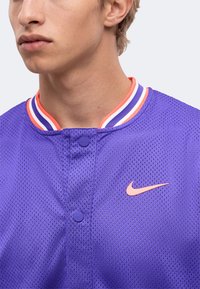Νέος που φοράει μωβ δίχτυ αθλητική φανέλα Nike με πολύχρωμο ριγέ γιακά και ροδακινί λογότυπο Nike στο στήθος.