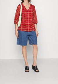 Blusa a cuadros rojos con escote en V, combinada con pantalones cortos de mezclilla. Mules de tacón bloque negro y un bolso de hombro verde claro.