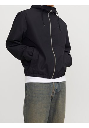 Person iført en sort lynlås hættejakke med hænderne i lommerne, hvid skjorte under og falmede blå jeans.